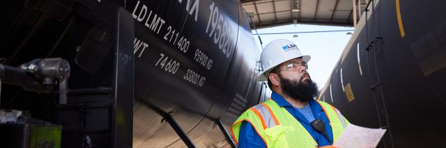 "Industrial worker wearing a hard hat and high-visibility safety vest reviewing paperwork between two black tanker railcars inside a covered rail loading facility."