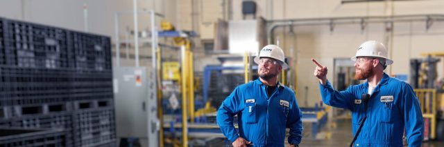 Two industrial workers in blue coveralls and hard hats standing on a factory floor, discussing work while one gestures toward equipment.